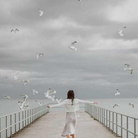 seagulls flying over a woman walking on a dock
